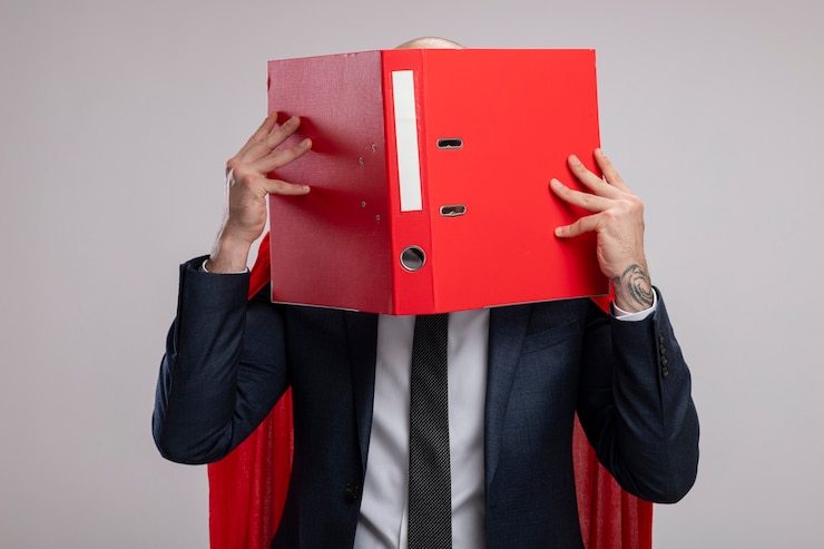 Free Photo super hero bearded business man in red cape holding open folder covering his face behind it standing over white wall